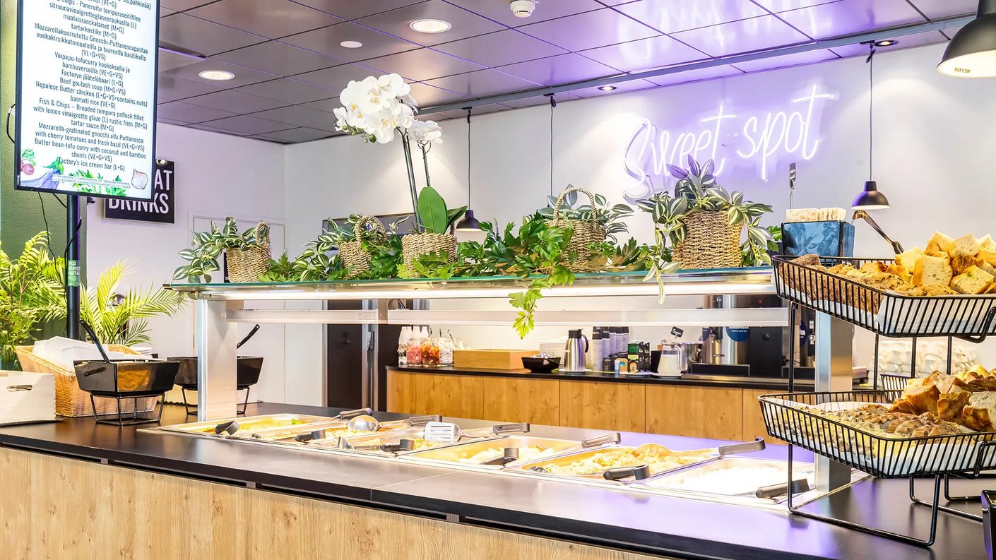Moderni buffet-ravintola, jossa on tarjolla lämpimiä ruokia metallisissa tarjoiluastioissa, leipäkoreja sekä koristekasveja buffet-tason päällä. Taustalla näkyy neonvalokyltti 'Sweet spot' sekä digitaalinen näyttö, jossa on päivän ruokalista.