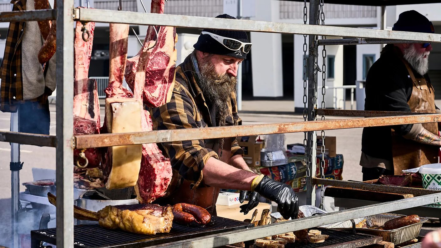 Mies laittaa ruokaa grillissä ulkona, taustalla näkyy lihaa roikkumassa ja muita lihoja valmiina syötäväksi. Hänellä on päässään musta pipo ja hänellä on sormikkaat käsissään. Taustalla on muita ihmisiä ja rakennuksia.