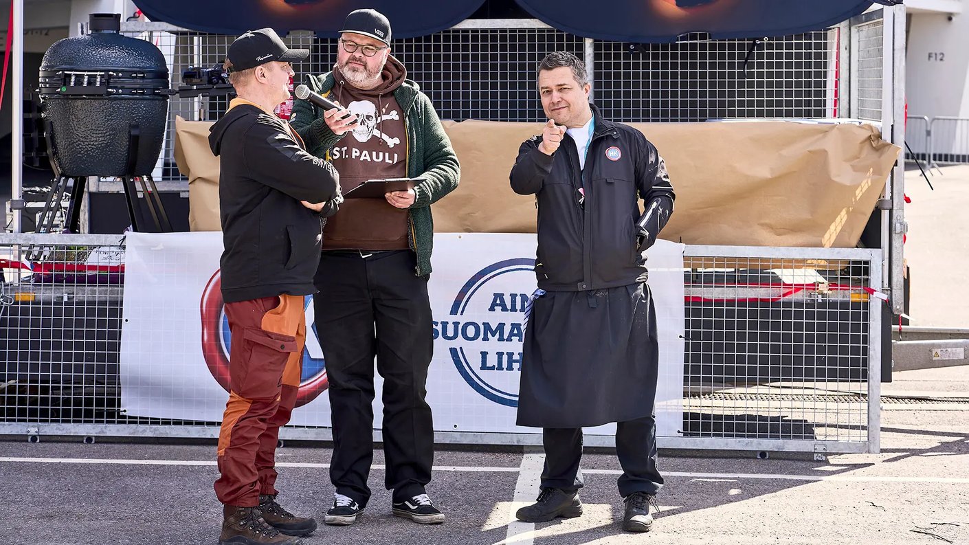 Kolme miestä keskustelee ja nauraa ulkona, takana on tapahtumakatos ja grillilaitteita. Mies vasemmalla puhuu ja pitää kädessään mikrofonia, keskellä oleva mies katsoo häntä ja pitää muistilistaa kädessään, ja oikealla oleva mies nyökkää ja viittaa sormellaan.