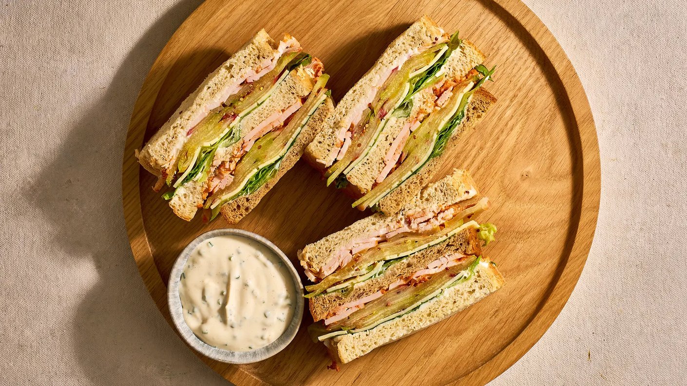 Puinen lautanen, jolla on neljä kolmioksi leikattua voileipää. Voileivissä on kerroksia, kuten kalkkunaa, salaattia, suolakurkkua ja majoneesia. Lautasen vieressä pieni kulho dippikastiketta. Tausta on vaalea kangasmainen pinta.