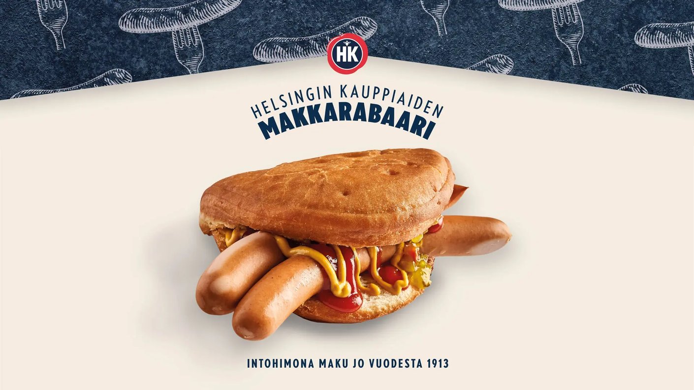 Mainoskuva HK Makkarabaarin lihapiirakasta kahdella nakilla vaalealla taustalla. Lihapiirakan välistä pilkottavat nakit on maustettu sinapilla, ketsupilla ja kurkkusalaatilla. Kuvan yläreunassa on sininen osio, jossa on makkara- ja haarukkapiirroksia, sekä HK-logo ja teksti ”Helsingin Kauppiaiden Makkarabaari”. Alareunassa lukee teksti ”Intohimona maku jo vuodesta 1913”.