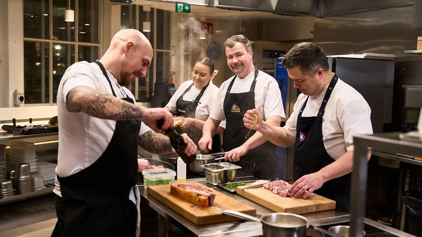 Neljä kokkia työskentelee vilkkaassa ammattikeittiössä ruostumattomasta teräksestä valmistetun työtason äärellä. Vasemmalla tatuoitu kokki levittää olutta lihaviipaleelle, kun taas oikealla kaksi kokkia leikkaavat ja valmistelevat raaka-aineita. Keskellä nainen ja viiksekäs mies kokki hymyilevät kameralle, kun mies sekoittaa kattilassa kiehuvaa ruokaa, josta nousee höyryä. Kaikilla kokkeilla on yllään valkoinen kokintakki ja musta esiliina, osassa esiliinoista on logoja. Taustalla näkyy keittiölaitteita, kuten uuneja ja hyllyjä, sekä ikkunoita. Valo on kirkas ja työtaso on täynnä työkaluja ja raaka-aineita.