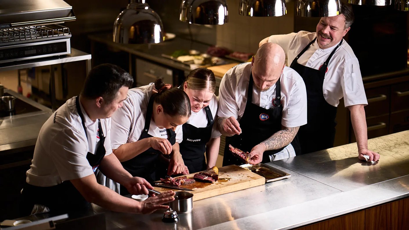 Yläviistosta otettu kuva viidestä kokista, jotka ovat kerääntyneet ruostumattomasta teräksestä valmistetun työpöydän ääreen ammattikeittiössä. Pöydällä on puisella leikkuulaudalla kypsennettyjä lihanpaloja, joita he tarkastelevat.  Keskellä oleva tatuoitu, kalju mies ja hänen vieressään oleva nainen sirottelevat sormisuolaa lihalle. Toinen mieskokki vasemmalla pitää pientä kattilaa, ja kolmas nainen tarkkailee lihan takaa. Oikealla oleva mieskokki katsoo hymyillen vasemmalle, käsi pöydällä. Kaikilla on yllään valkoinen kokintakki ja musta esiliina, joissa on HK-logoja. Taustalla näkyy keittiölaitteita ja lämpölamppuja, ja valaistus on pistemäinen ja lämmin.