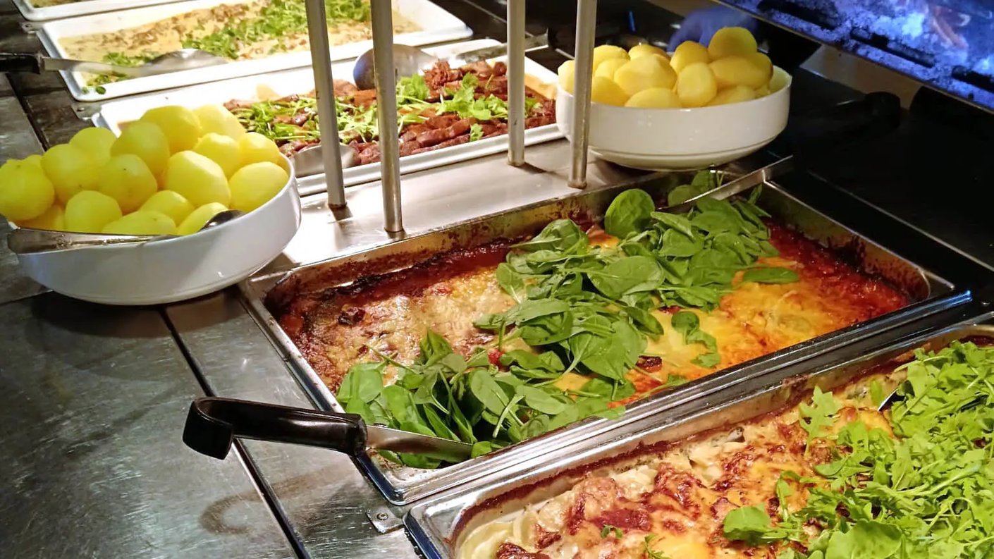 Lähikuva buffet-pöydän tarjoiluastiasta, joka on täynnä lämpimiä ruokia. Etualalla on kaksi GN-astiaa, joissa on paistettuja ruokia, joista päällimmäinen on koristeltu tuoreilla pinaatinlehdillä. Sen vieressä ja takana näkyy kulhoissa tarjoiltavia keitettyjä perunoita.