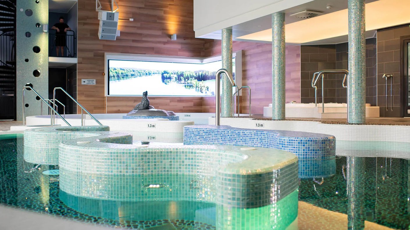 matran kylpylän sisätiloissa otettu kuva "Taika-altaasta" (Magic Pool), joka on moderni allasalue turkoosein mosaiikkilaatoin. Allas on monimuotoinen, ja sen keskellä on pieni patsas. Taustalla on suuri digitaalinen näyttö, joka esittää maisemaa Saimaan vesistöstä. 