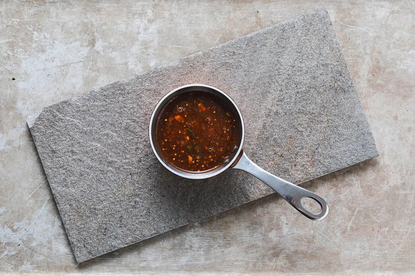 Pieni teräskattila kivialustalla ylhäältä päin kuvattuna, sisällä tummaa sinapinsiemeniä sisältävää kastiketta.