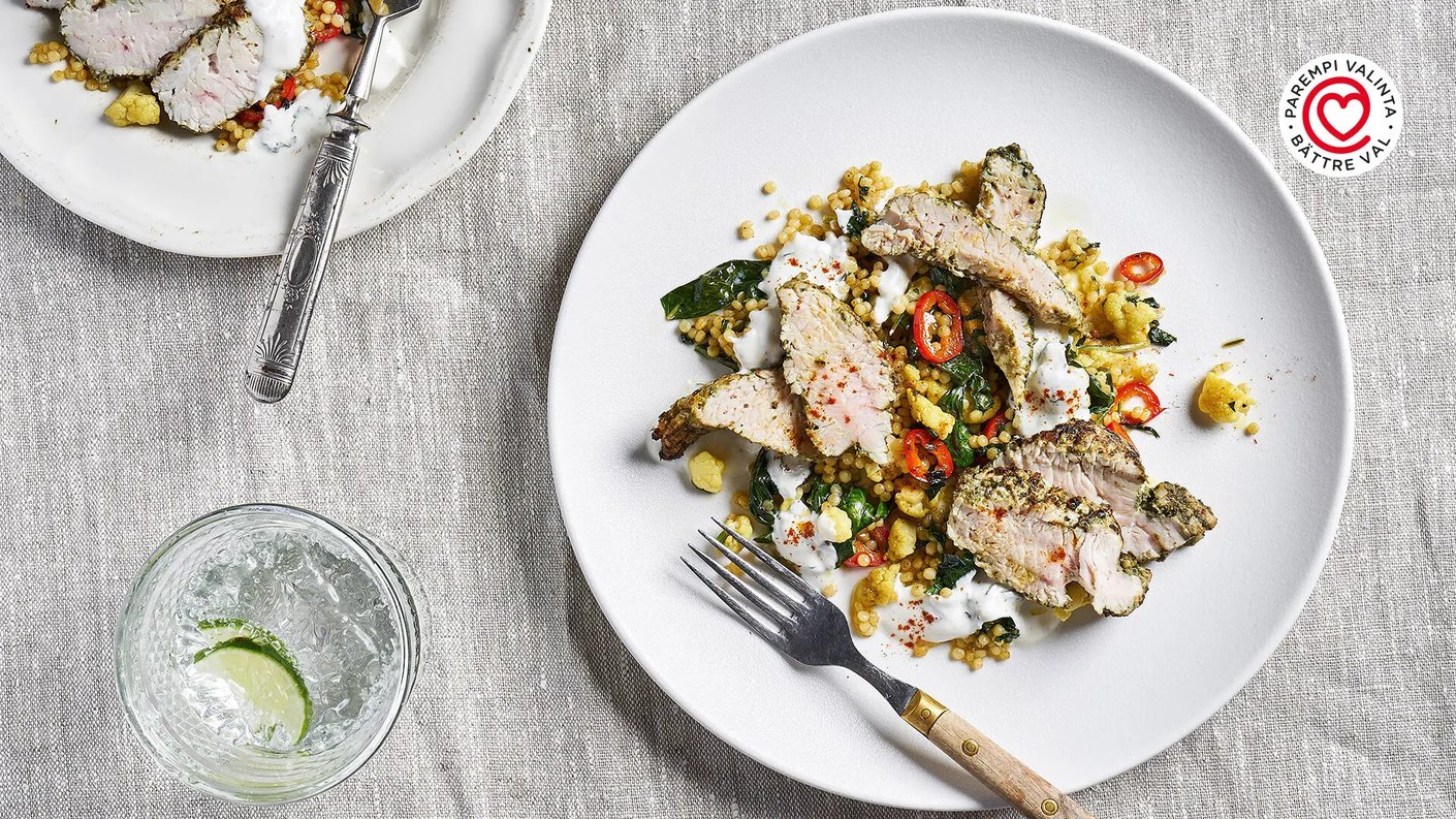  Yläkuva modernista kalkkunan sisäfileeateriasta valkoisella lautasella. Kalkkuna on viipaloitu ja maustettu vihreällä pestolla tai yrteillä, ja se on asetettu kuskusin tai vastaavan viljan, vihreiden lehtien, ja keltaisten kasvisten pedille. Annos on koristeltu punaisilla chilirenkailla ja valkoisella kastikkeella. Oikeassa yläkulmassa on punavalkoinen logo, jossa lukee "PAREMPI VALINTA • BÄTTRE VAL". Vasemmalla näkyy juomalasi, jossa on kirkasta juomaa, jäitä ja limelohko. 