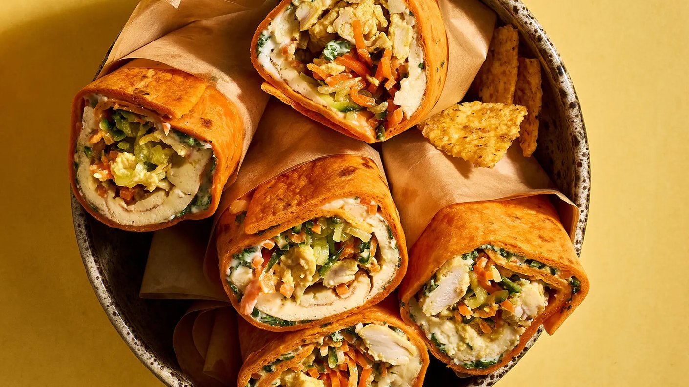 Annos tortillawrappeja, joissa täytteenä Kariniemen Kananpoikaa, kasviksia, kuten porkkanaa ja salaattia, sekä kastiketta. Wrapit on kääritty oranssiin tortillapohjaan ja aseteltu kulhoon. Kulhossa näkyy myös muutama nacho. Tausta on keltainen.