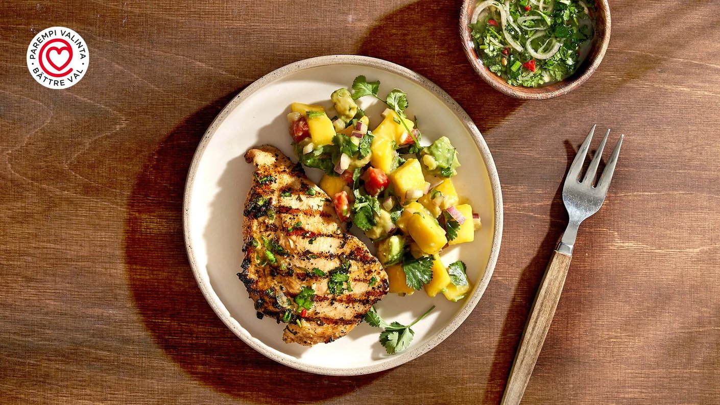 Ylhäältä otettu kuva lautasesta, jossa on grillattu, maustettu karibian jerk -minuuttipihvi broilerista, jolla on selkeät grillausraidat. Pihvin vieressä on kirkkaan keltainen ja vihreä mango-salsa tai hedelmäinen lisuke. Annos on asetettu puupöydälle. Vasemmassa yläkulmassa on punavalkoinen "PAREMPI VALINTA • BÄTTRE VAL" -logo.