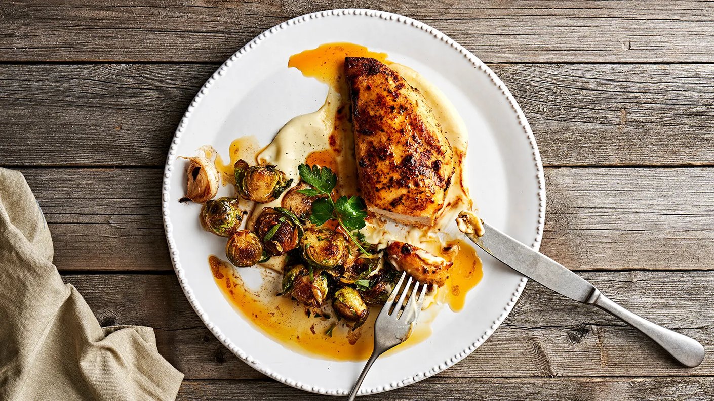 Annos, jossa on paistettua kanaa, paahdettuja ruusukaaleja ja pyreetä. Annos on pyöreällä, valkoisella lautasella. Lautasella on myös haarukka ja veitsi. Taustalla on tumma, puinen pöytä, ja lautasen vieressä on pellava- tai puuvillaliina.