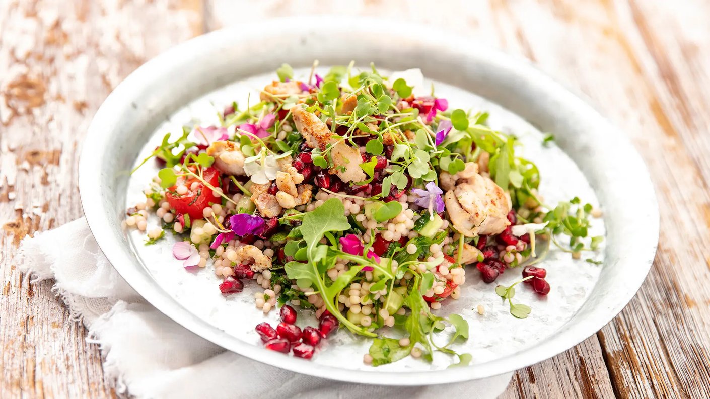 Monivärinen marokkolaistyylinen kanasalaatti, jossa on couscousia, granaattiomenan siemeniä, lehtivihanneksia ja syötäviä kukkia, on esillä pyöreällä, metallisella lautasella. Lautasen alla on valkoinen lautasliina.