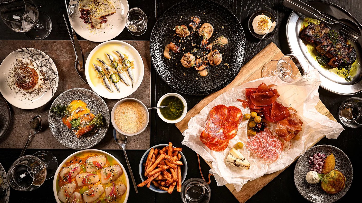 Pöydällä on pieniä lautasia, joissa on erilaisia välimerellisiä tapaksia. Lisäksi kuvassa on leikkuulauta, jossa on erilaisia leikkeleitä, oliiveja sekä juuston paloja. Kuvan oikeassa yläkulmassa on grillattua lihaa teräsvadilla ja oikeassa alakulmassa bliniannos. Tyhjät viinilasit ympäröivät ruokaa.