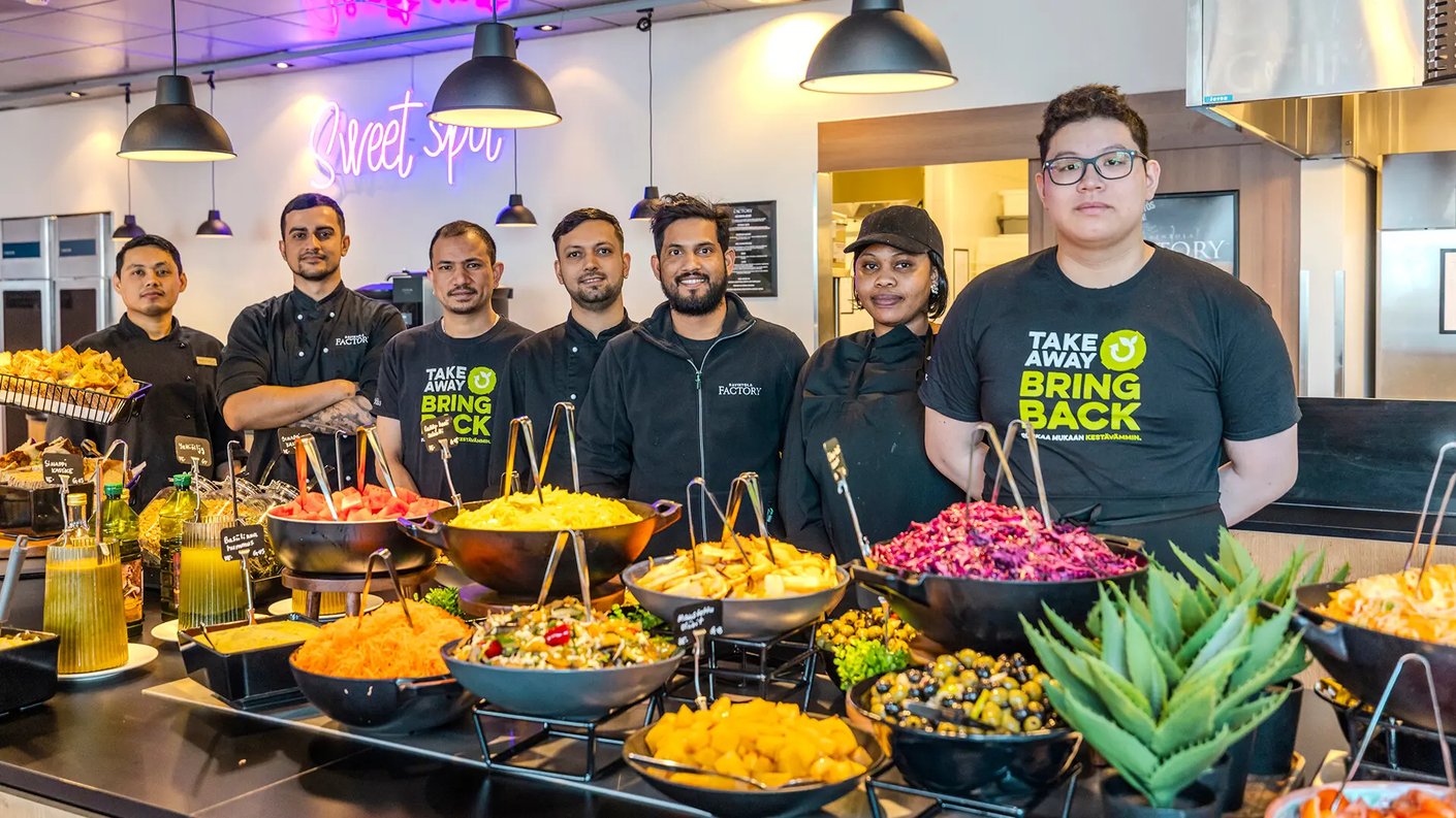 Ravintolan henkilökunta seisoo buffetpöydän takana, joka on täynnä värikkäitä salaatteja ja muita ruokia. Taustalla neonvaloissa teksti 'Sweet spot' ja moderni sisustus. Henkilökunnalla on ravintola- ja kampanjapaitoja yllään.