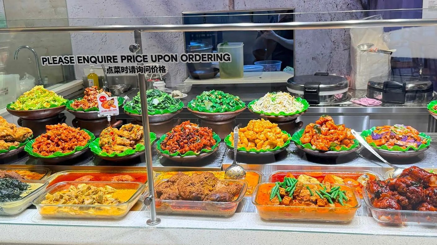 Näkymä aasialaisen ravintolan noutoruokatiskiin, jossa on esillä runsas valikoima valmiita ruokalajeja kolmessa tasossa.      Ylhäällä: Erilaisia paistettuja vihanneksia, kuten kaalia, ituja ja lehtivihreitä saviruukuissa.      Keskellä: Valikoima friteerattuja ja kastikkeessa olevia liharuokia, pääasiassa kanaa ja porsasta.      Alhaalla: Lasivuokiin aseteltuja haudutettuja curryja, kookospohjaisia kastikkeita ja paahdettuja kanan koipia.      Yksityiskohta: Lasitiskin yläreunassa on kyltti, jossa pyydetään tiedustelemaan annosten hintaa tilattaessa.