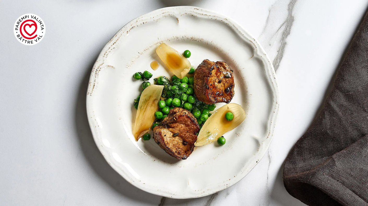 Lähikuva valkoisella, karkeareunaisella lautasella olevasta ateriasta. Lautasella on kaksi pyöreää, paistettua, maustettua lihanoiduuttia, jotka on asetettu kasan päälle höyrytettyjä herneitä. Herneiden joukossa on muutama lohko vaaleaa, paistettua tai haudutettua fenkolia tai sipulia, jotka on valeltu kirkkaalla kastikkeella. Annos on asetettu marmorisen näköisen pinnan päälle. Kuvan vasemmassa yläkulmassa on pyöreä punavalkoinen logo, jossa lukee "PAREMPI VALINTA • BÄTTRE VAL".