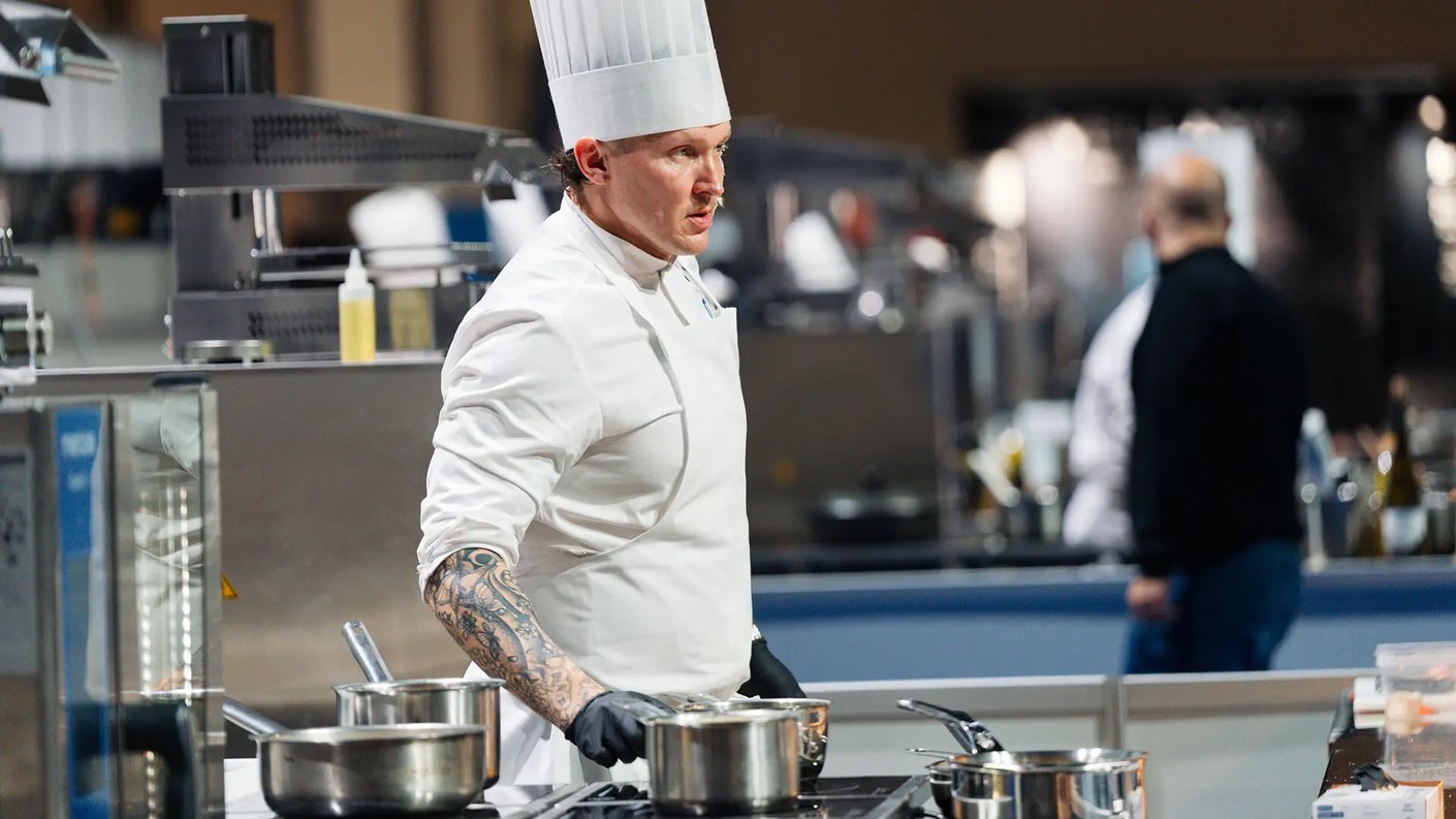 Vuoden Kokki Simo Harrivaara työskentelemässä keittiössä, pukeutuneena valkoiseen kokin takkiin ja korkeaan kokin hattuun, keskittyen ruokien valmistukseen ruostumattomien teräskattiloiden äärellä. Hänen oikeassa käsivarressaan näkyy tatuointeja ja hän käyttää mustia hansikkaita. Taustalla näkyy keittiövälineitä ja hämärästi valaistuja hahmoja. Oikeassa yläkulmassa on Vuoden Kokki -logo.