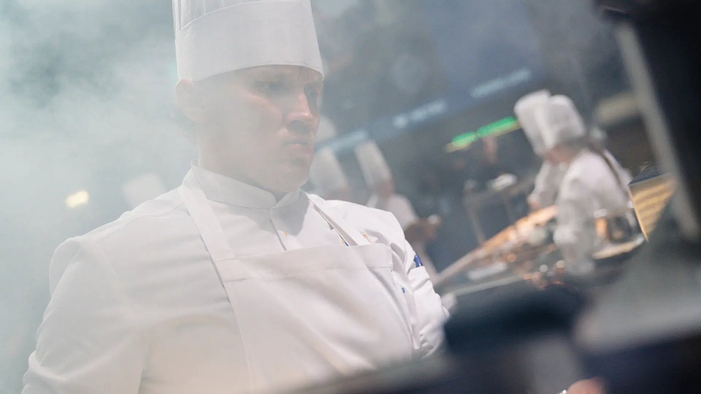 Vuoden Kokki Simo Harrivaara näkyy intensiivisesti keskittyneenä, pukeutuneena valkoiseen kokin takkiin ja korkeaan kokin hattuun. Kuvassa on savua, joka luo dramaattisen tunnelman hänen ympärilleen. Taustalla näkyy muita kokkeja työskentelemässä keittiössä.