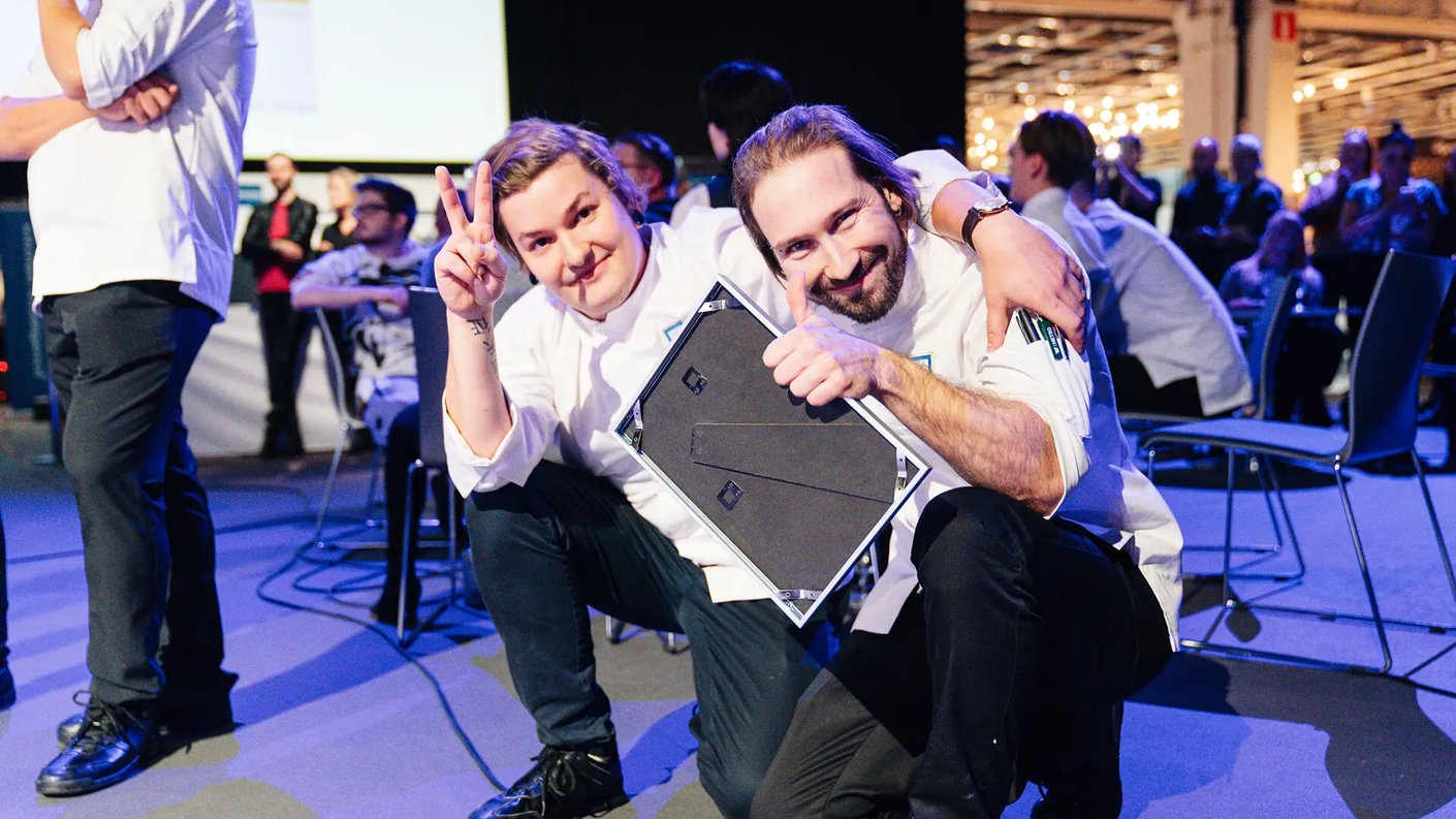 Kaksi hymyilevää Vuoden Kokki 2023 kisan henkilöä kokkitakeissa poseeraa lähekkäin kyykkyasennossa lavan edustalla. Toinen heistä näyttää rauhanmerkkiä käsillään ja toinen pitää kädessään kehystettyä palkintoa tai diplomia samalla peukuttaen. Taustalla näkyy muita ihmisiä istumassa ja seisomassa tapahtumatilassa, jossa on valaistuja koristevaloja ja avointa tilaa.