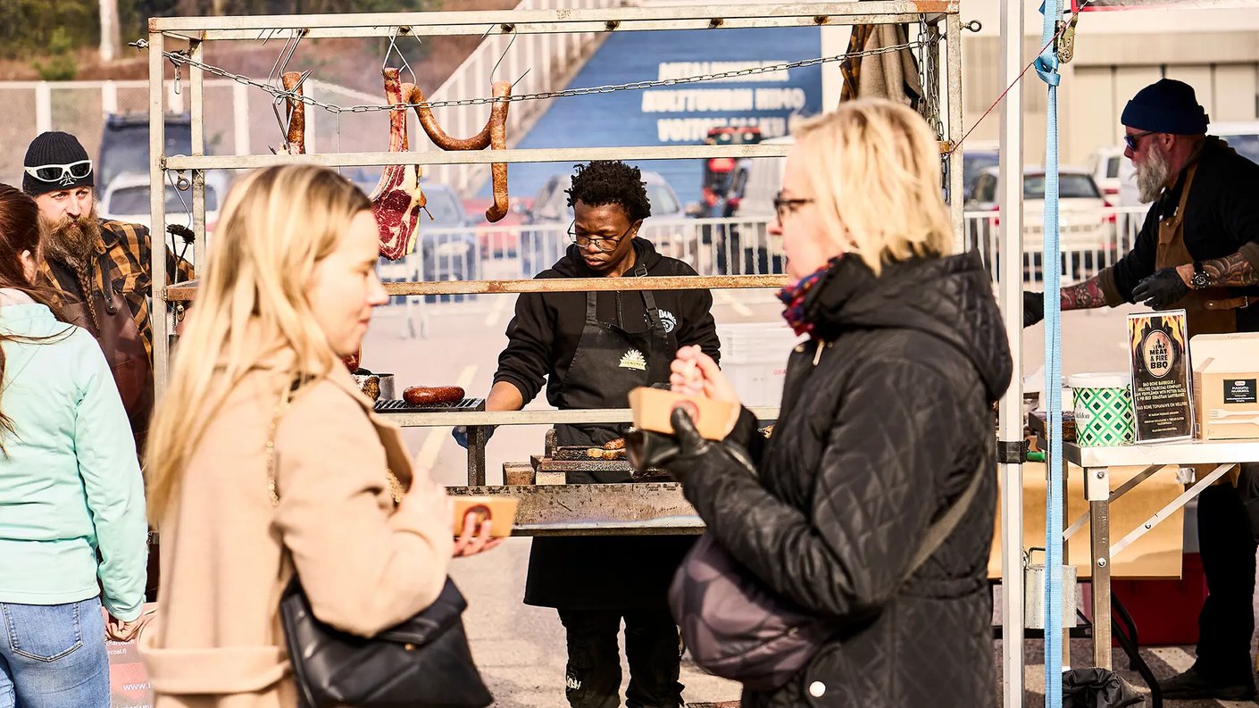 Ulkoilmatapahtuma Meat&Fire BBQ, jossa ihmiset jonottavat ja nauttivat grilliruoasta. Kuvassa näkyy grillikatos, jossa lihaa paistetaan avotulella. Keskellä grillaa mustaan essuun pukeutunut henkilö, taustalla on muita myyjiä ja asiakkaita. Etualalla kaksi naista syö grilliruokaa pahvilaatikoista. Tapahtuma vaikuttaa vilkkaalta ja aurinkoiselta.
