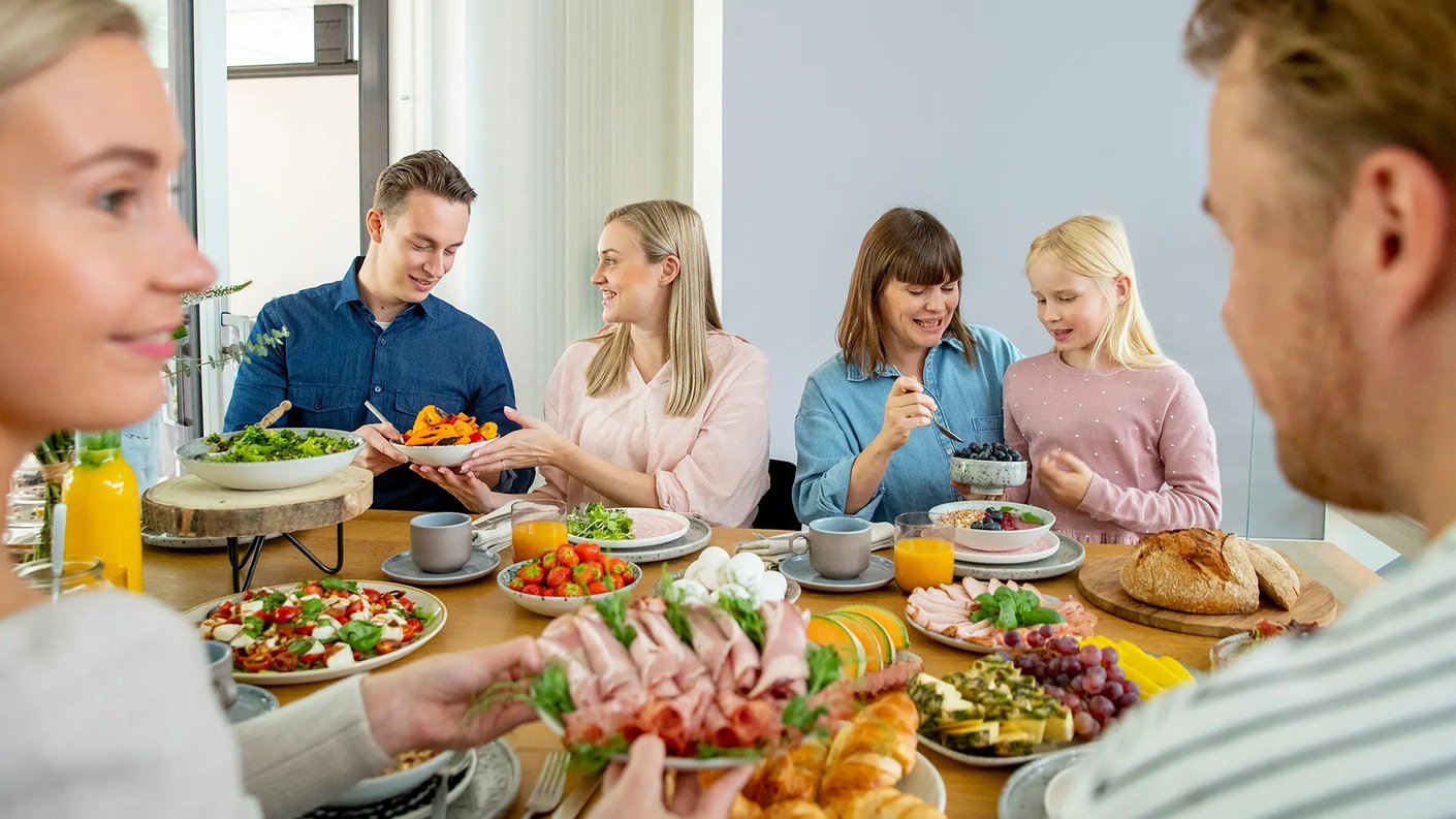 Iloisia ihmisiä brunssilla. Pöydässä leikkeleitä, leipää, hedelmiä, marjoja ja vihanneksia.
