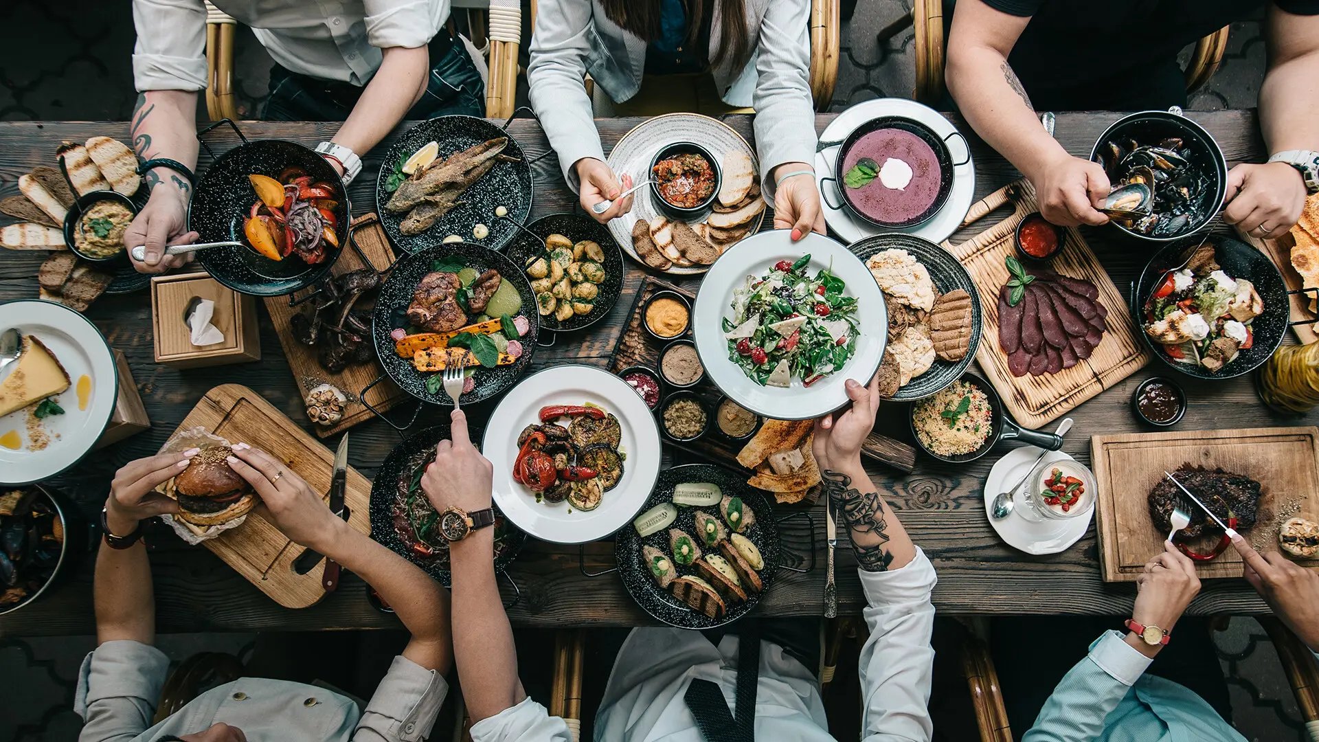 Ruokapöydällä runsaasti erilaisia jaettavia ruokia astioissa. Pöydän molemmin puolin ihmisiä syömässä.
