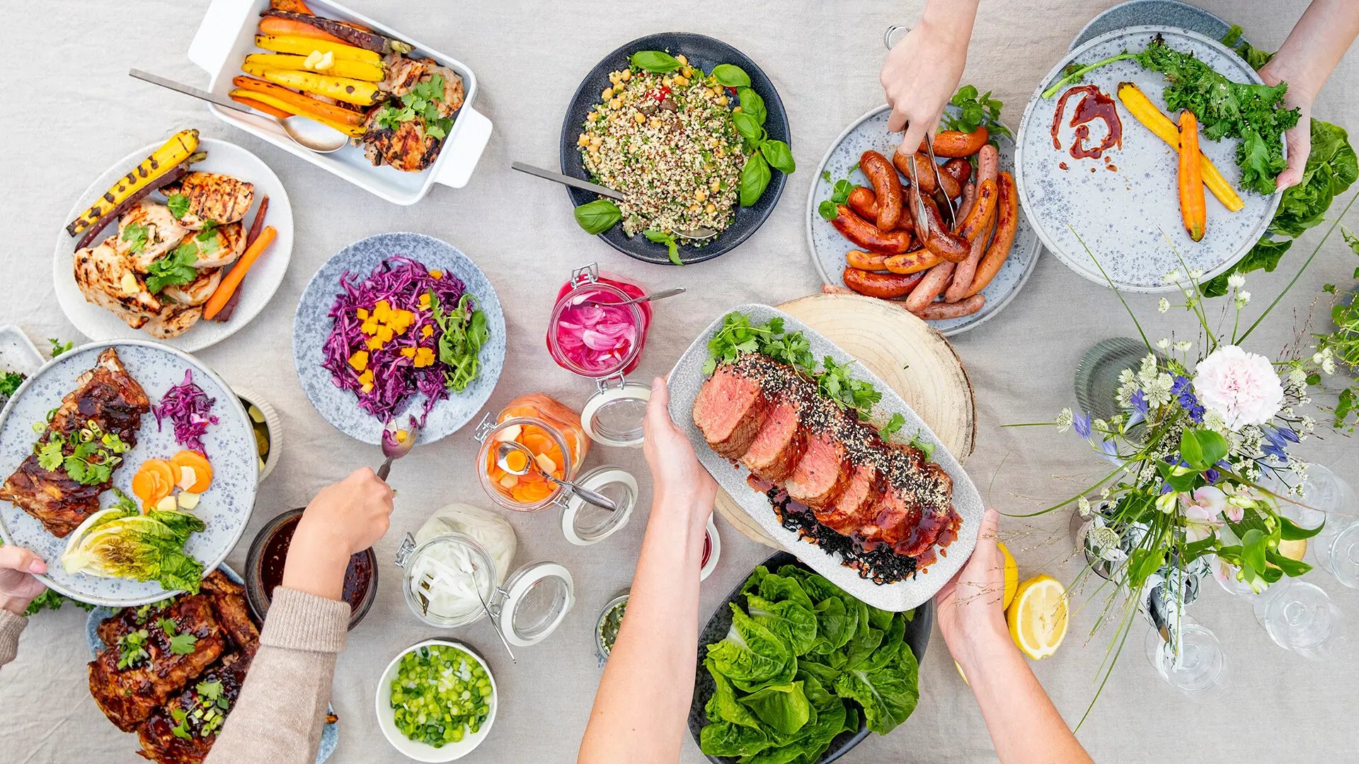 Kesäinen ruokapöytä, jossa on monipuolinen valikoima grilliruokia ja lisukkeita. Näkyy grillattuja kasviksia, makkaraa, lihaa, salaattia, punakaalisalaattia ja marinoituja vihanneksia. Koristeluna kukka-asetelma ja muita kesäisiä yksityiskohtia. Kädet tarjoilevat annoksia pöydästä.