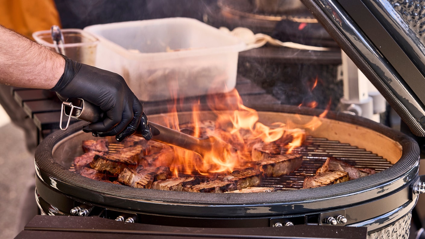 Mies kumihanskat kädessään grillaa lihaa grillissä, jossa on palavia hiiliä ja pihviä. Taustalla näkyy muovinen astia ja osittain auki oleva grillin kansi.