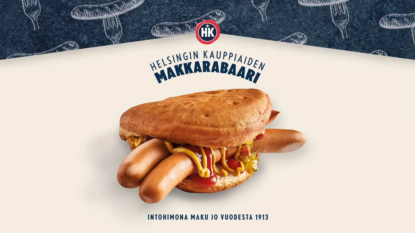 Mainoskuva HK Makkarabaarin lihapiirakasta kahdella nakilla vaalealla taustalla. Lihapiirakan välistä pilkottavat nakit on maustettu sinapilla, ketsupilla ja kurkkusalaatilla. Kuvan yläreunassa on sininen osio, jossa on makkara- ja haarukkapiirroksia, sekä HK-logo ja teksti ”Helsingin Kauppiaiden Makkarabaari”. Alareunassa lukee teksti ”Intohimona maku jo vuodesta 1913”.