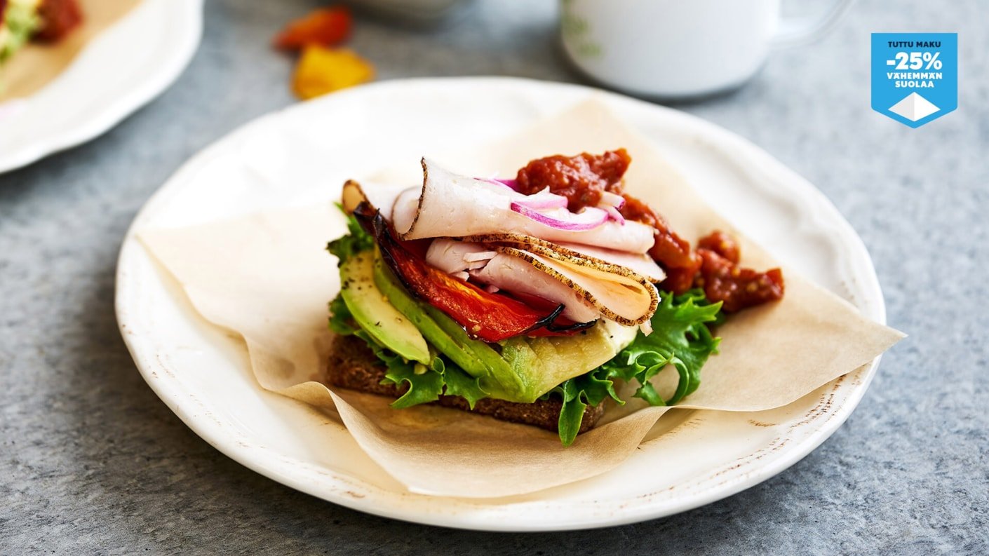 Lähikuva avoimesta ruisleipäsandwichista pyöreällä, valkoisella lautasella. Leivän päällä on kerroksittain vihreää salaattia, viipaloitua avokadoa, paahdettua tomaattia ja päällimmäisenä siivuja kanaleikettä tai kalkkunaa, joka on koristeltu punasipulirenkailla ja tummalla kastikkeella/salsalla. Leipä on asetettu lautaselle leivinpaperin palan päälle. Kuvan oikeassa yläkulmassa on sinivalkoinen logo, jossa lukee "TUTTU MAKU –25 % VÄHEMMÄN SUOLAA".