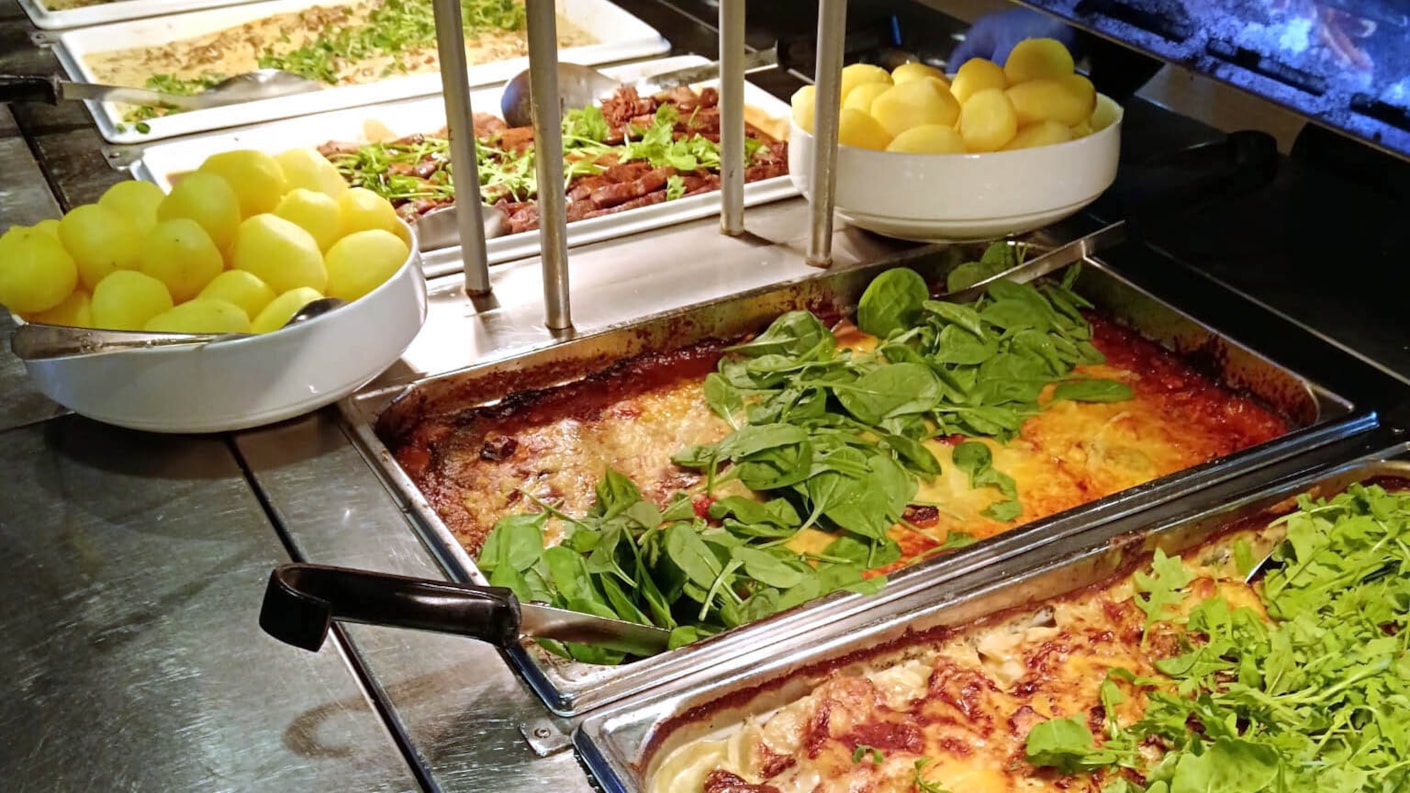 Lähikuva buffet-pöydän tarjoiluastiasta, joka on täynnä lämpimiä ruokia. Etualalla on kaksi GN-astiaa, joissa on paistettuja ruokia, joista päällimmäinen on koristeltu tuoreilla pinaatinlehdillä. Sen vieressä ja takana näkyy kulhoissa tarjoiltavia keitettyjä perunoita.