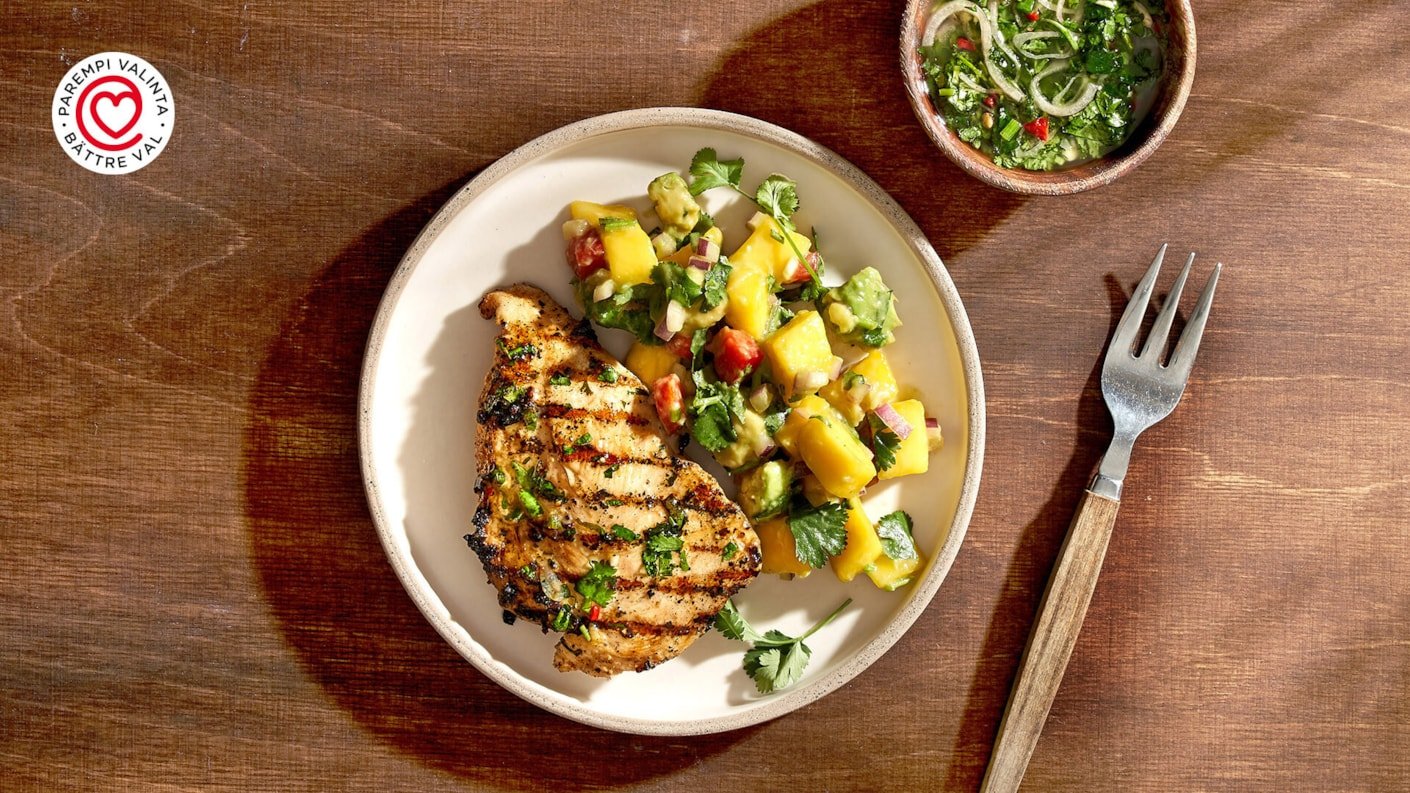 Ylhäältä otettu kuva lautasesta, jossa on grillattu, maustettu karibian jerk -minuuttipihvi broilerista, jolla on selkeät grillausraidat. Pihvin vieressä on kirkkaan keltainen ja vihreä mango-salsa tai hedelmäinen lisuke. Annos on asetettu puupöydälle. Vasemmassa yläkulmassa on punavalkoinen "PAREMPI VALINTA • BÄTTRE VAL" -logo.