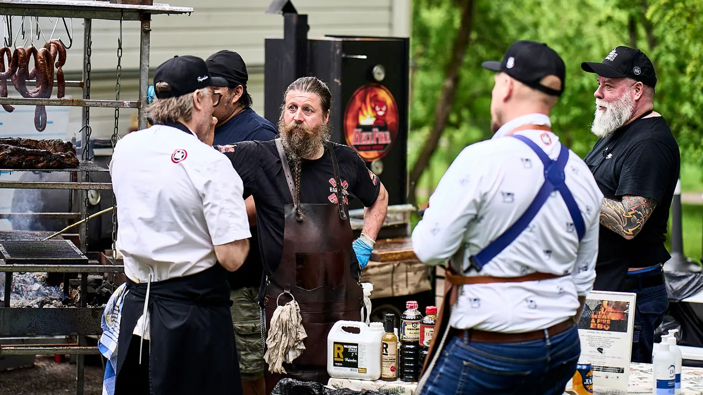 Ryhmä kokkeja ja grillimestareita keskustelee ulkona BBQ-tapahtumassa suuren grilli- ja savustuslaitteiston äärellä. Vasemmalla riippuu makkaroita, ja pöydällä etualalla on erilaisia kastike- ja maustepulloja. Keskellä seisoo pitkäpartainen mies nahkaisessa esiliinassa katsoen puhujaa.